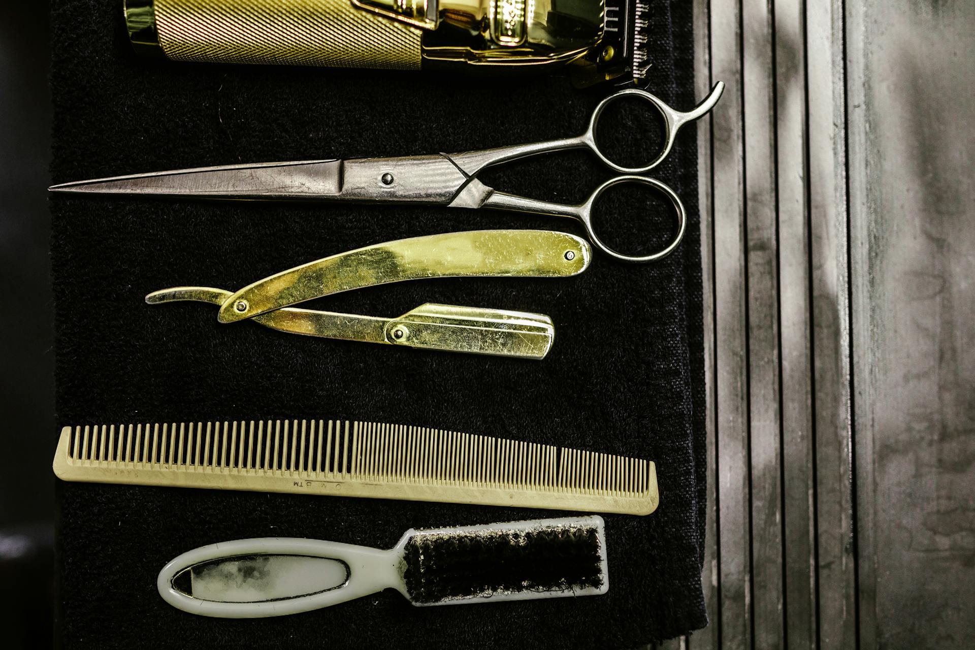 Flatlay of gold clipper, scissors, straight razor, comb and brush on dark cloth